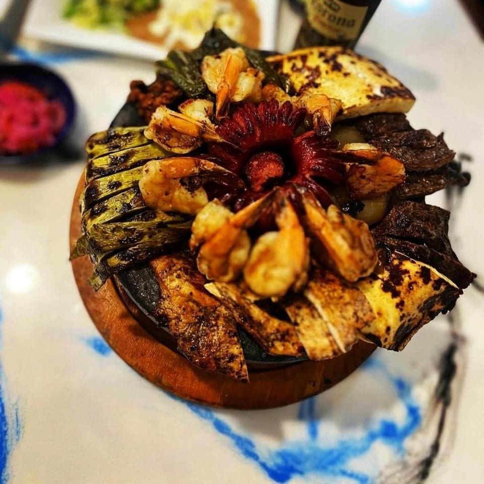 A sizzling platter of grilled shrimp, steak, cheese, and vegetables, with a red flower garnish.