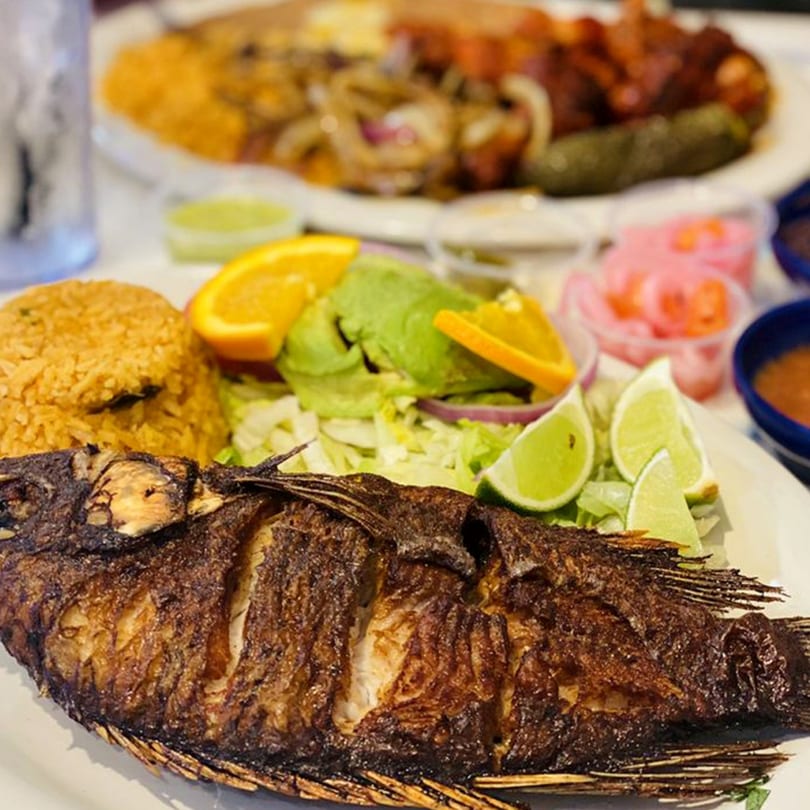 A whole fried fish with rice, salad, orange slices, and lime wedges on a white plate.
