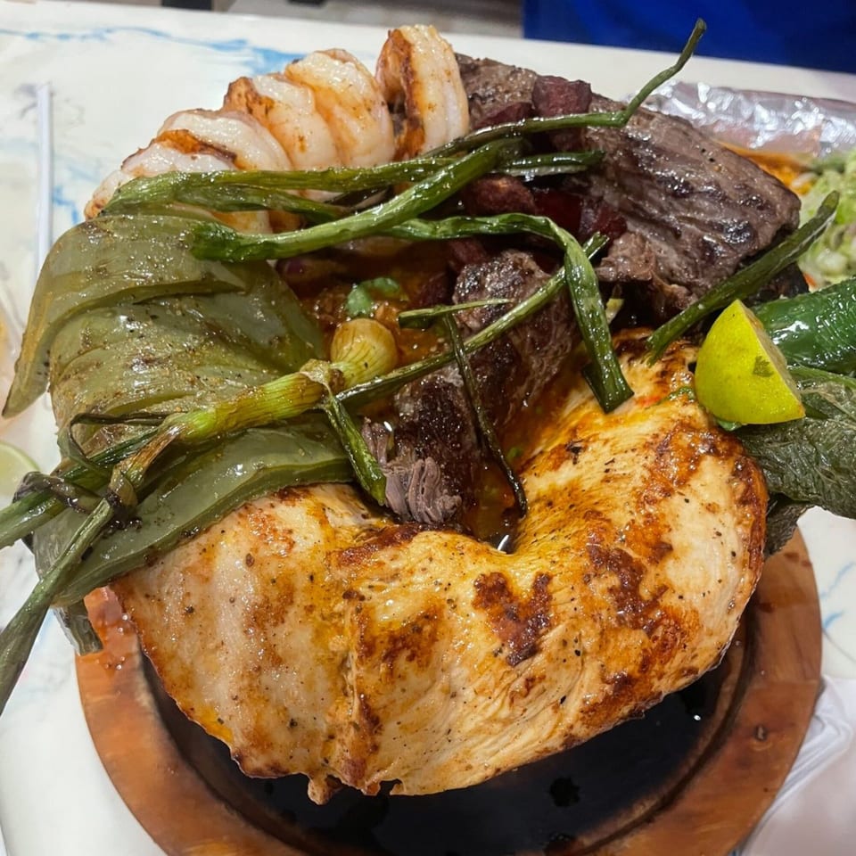 A sizzling Mexican molcajete dish with grilled chicken, steak, shrimp, green onions, and lime.