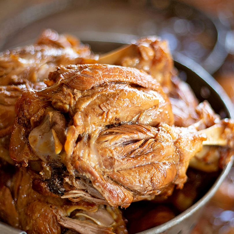 Close-up of tender, braised Mexican pork shank, glistening with sauce in a metal pot.